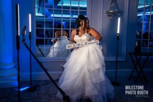 A bride in a white wedding gown posing with a "Happily Ever After" sign in a photo booth setup at Milestone NJ.