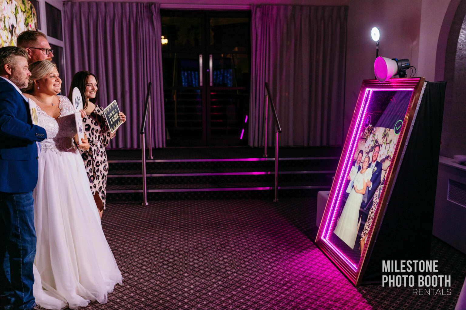 A sleek, modern mirror booth with glowing neon lights reflects a luxurious event space, with guests in the background waiting for their turn. - Milestone Photo Booth