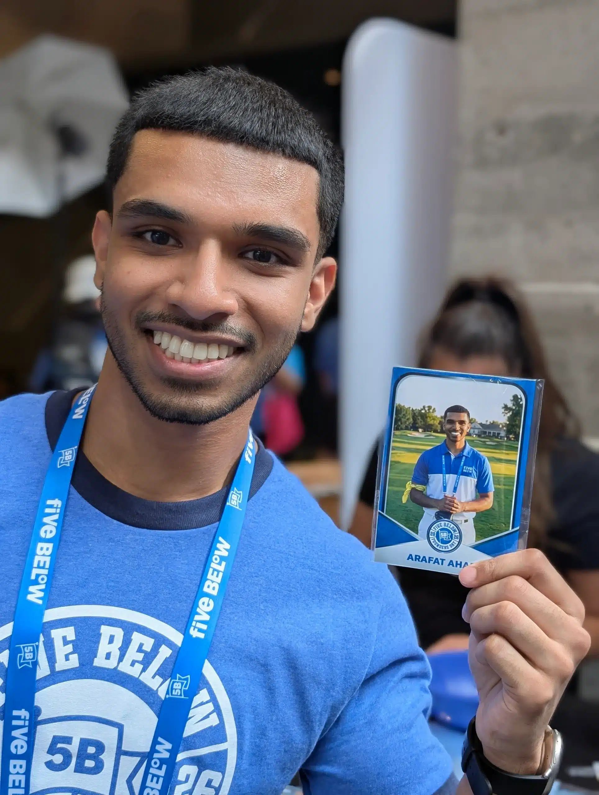 A Five Below employee holds up his custom AI trading card at Founders Day 2026, showing him as a golfer in Five Below branded gear
