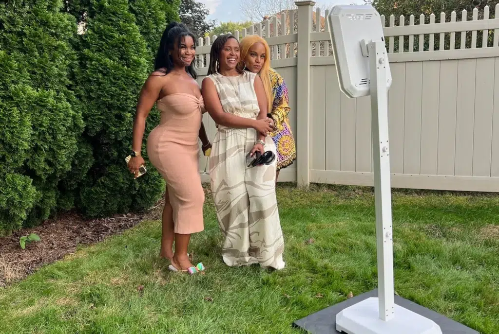 Three guests in dressy outfits posing beside a white DIY photo booth on a stand in a grassy backyard with a tan picket fence