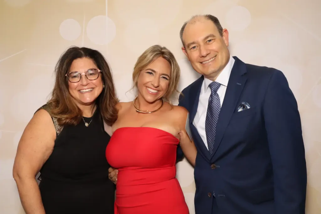 Three guests in formal attire posing together in front of the gold bokeh backdrop at the AAML New Jersey installation gala