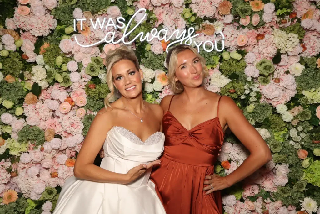 Bride and bridesmaid pose in front of a pink flower wall with a neon sign reading It was always you at a Crystal Springs Resort wedding