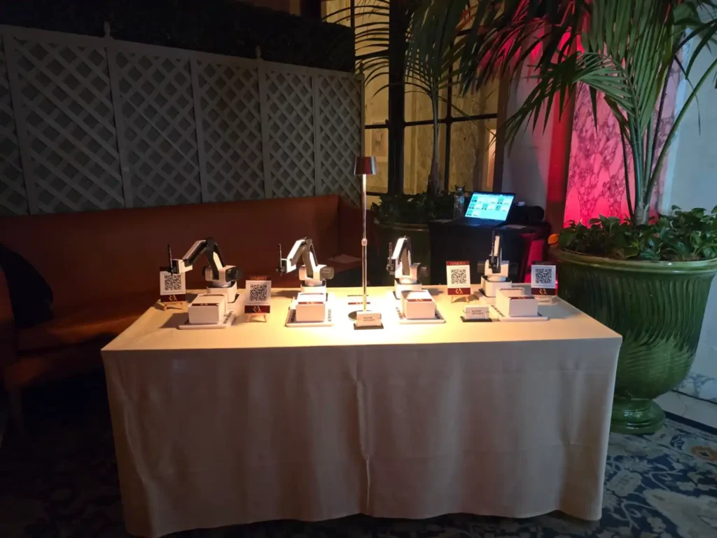 Four AI sketch robots at The Plaza Hotel on a draped table with custom RA monogram signage, surrounded by palms and warm uplighting.